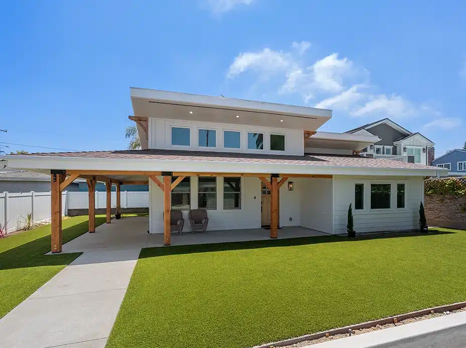 Modern two-story house with wide front porch, wood accents, white exterior, large windows, and manicured green lawn under blue sky.