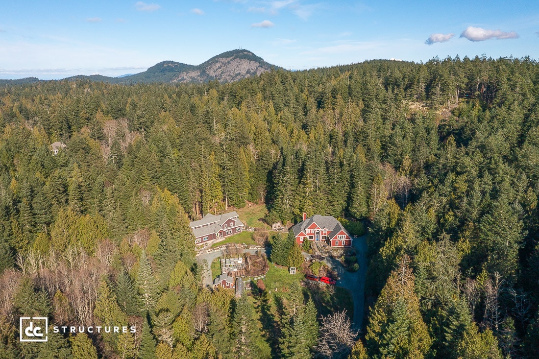 Aerial view of two large houses in dense evergreen forest with mountain backdrop and blue sky, connected by driveway and garden.