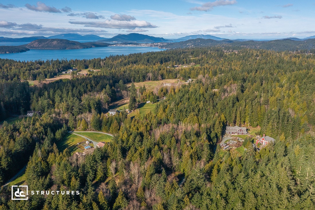 Aerial view of a forested rural landscape with scattered homes, open fields, and distant mountains near a large body of water under a partly cloudy blue sky.