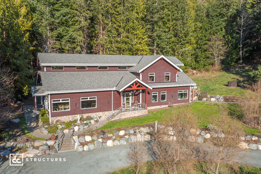 A large two-story house with a dark red exterior, gray roof, and stone accents, surrounded by trees and landscaping, seen from above.