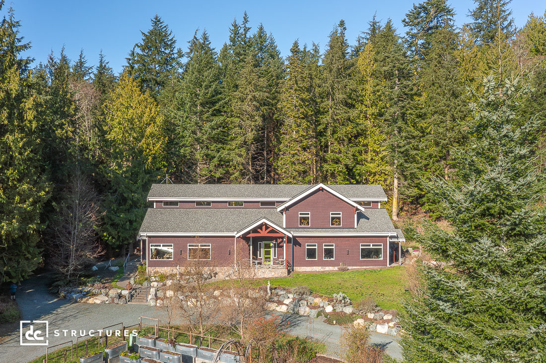 A large, two-story wood-framed house with a gabled roof sits among tall evergreens and landscaped greenery on a sunny day.