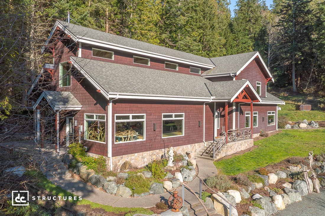 A two-story house with red siding, large windows, and stone foundation sits on a landscaped yard with trees, rocks, and a small porch.