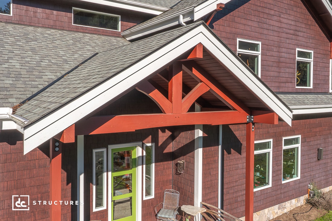 A close-up view of a house’s front entrance with a green door, red timber frame gable, brown shingle siding, porch chairs and table.