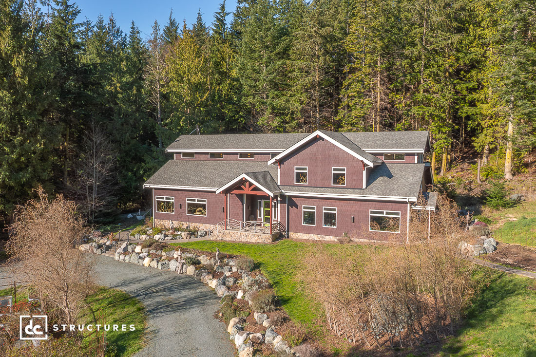 A large two-story maroon house with a gray roof sits in a forest clearing among pine trees, with a gravel driveway to the entrance.