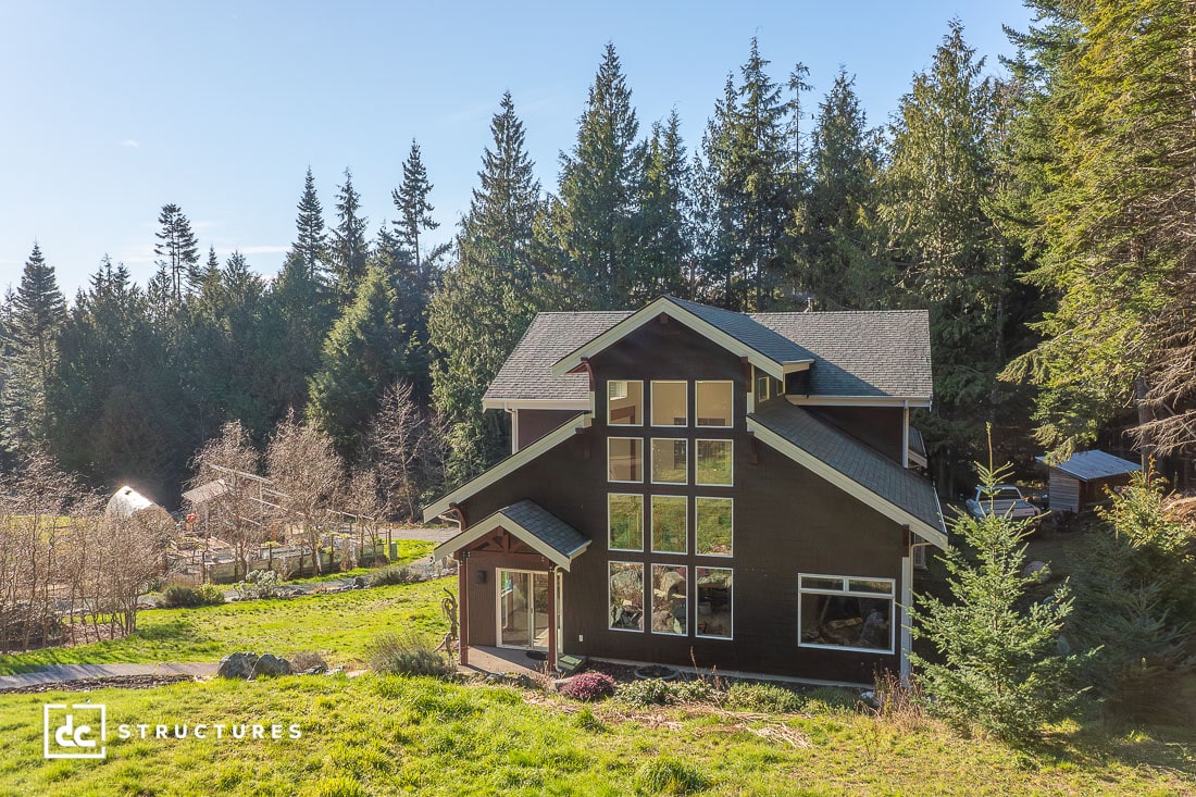 A modern two-story house with large windows is nestled among tall evergreens and greenery, featuring dark siding and a peaceful setting.