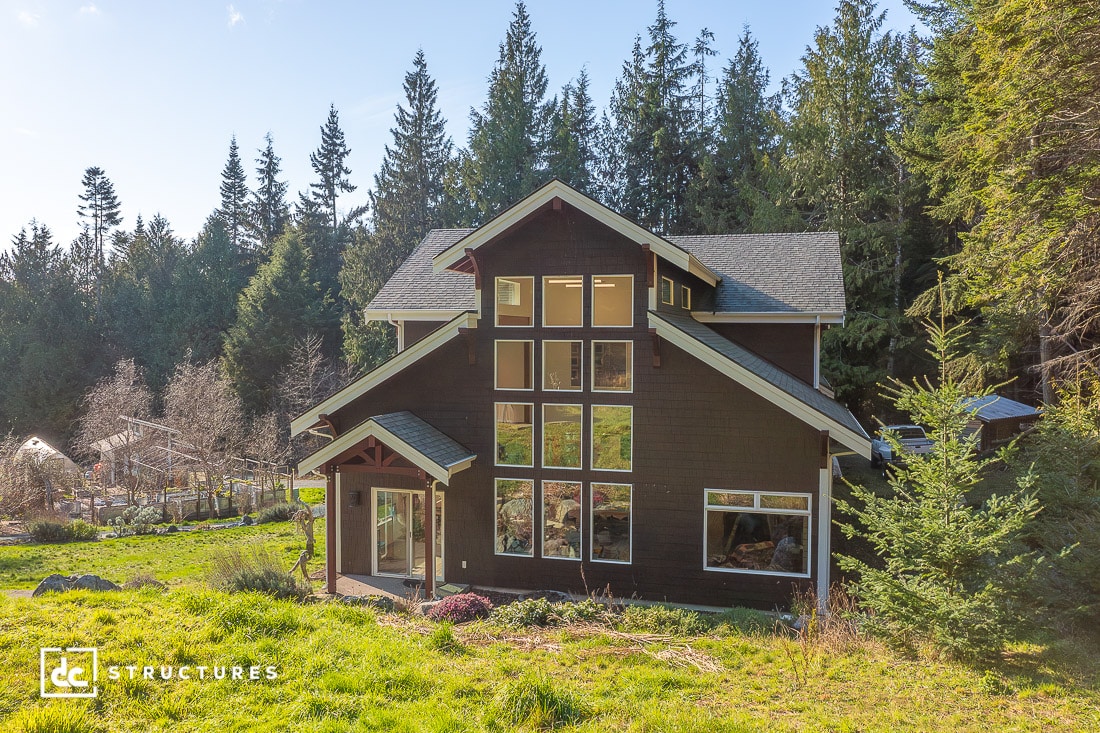 A two-story dark brown house with large, grid-like windows is surrounded by green grass and tall trees under a clear blue sky.