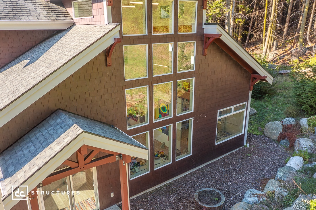 Aerial view of a modern house with large grid windows, brown siding, and gray shingle roof, surrounded by trees and a small garden.