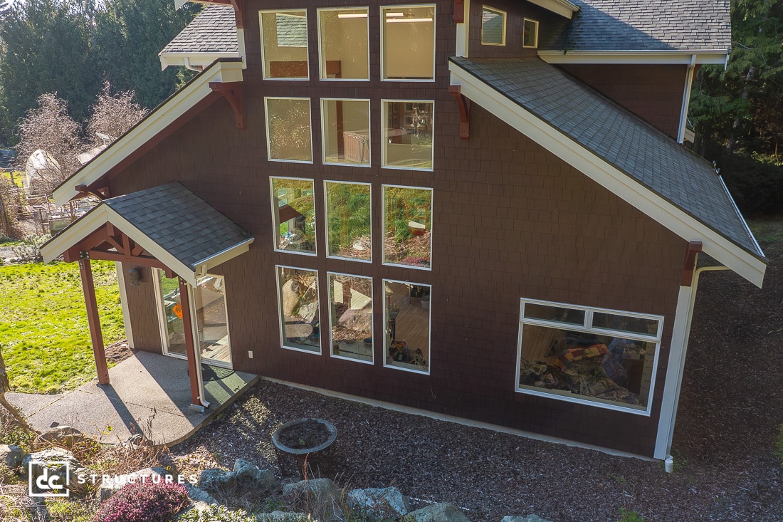A two-story house with large floor-to-ceiling windows and a brown exterior sits beside a garden and trees. Sunlight reflects off the windows, revealing a cozy interior.
