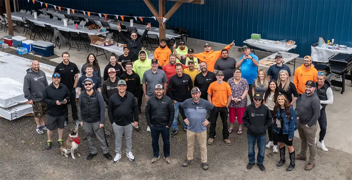 A large group of people pose and smile for a photo at an outdoor gathering with tables, tents, and food in the background. Some wear matching shirts, and one dog sits in front.