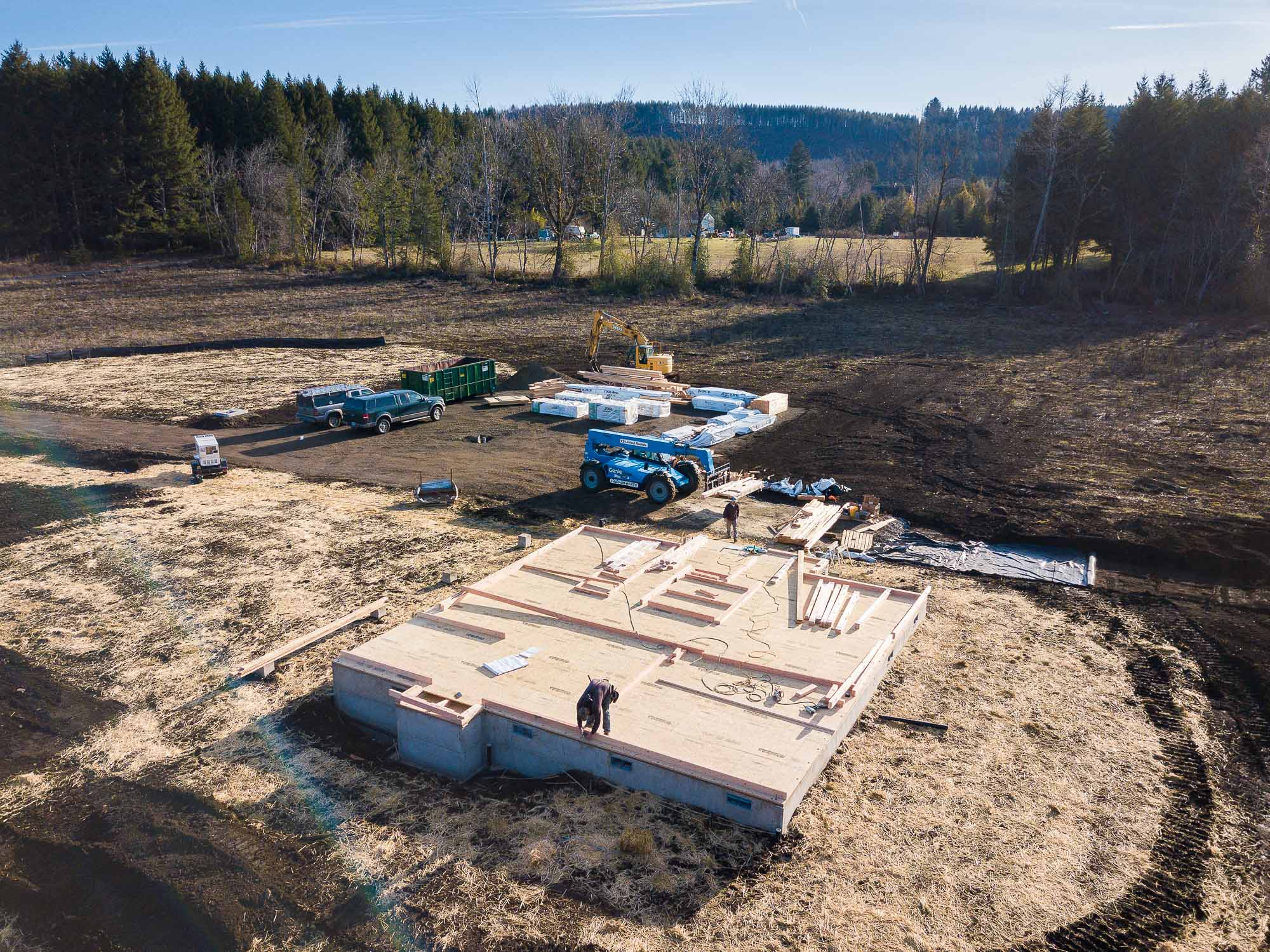 An aerial view of an outdoor living construction site featuring a concrete foundation with wooden floor joists, surrounded by equipment, building materials, and two construction workers.