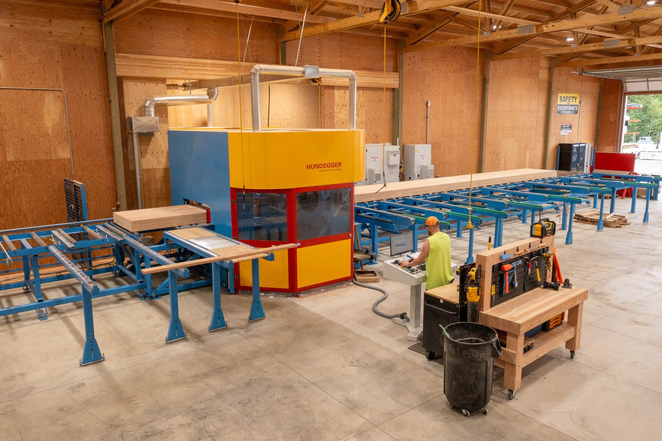 A worker in a high-visibility vest operates a computer at an industrial wood-processing machine, preparing materials for outdoor living.