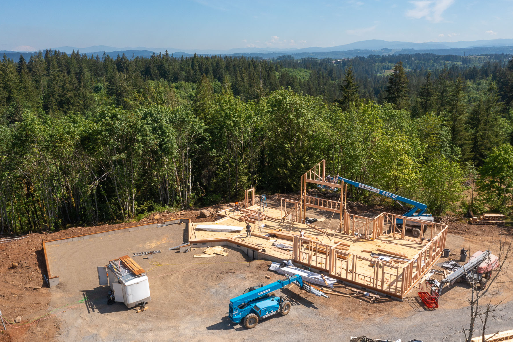 Aerial view of a wood-framed house under construction in a forest clearing, designed for outdoor living, with a crane and a utility trailer on site.
